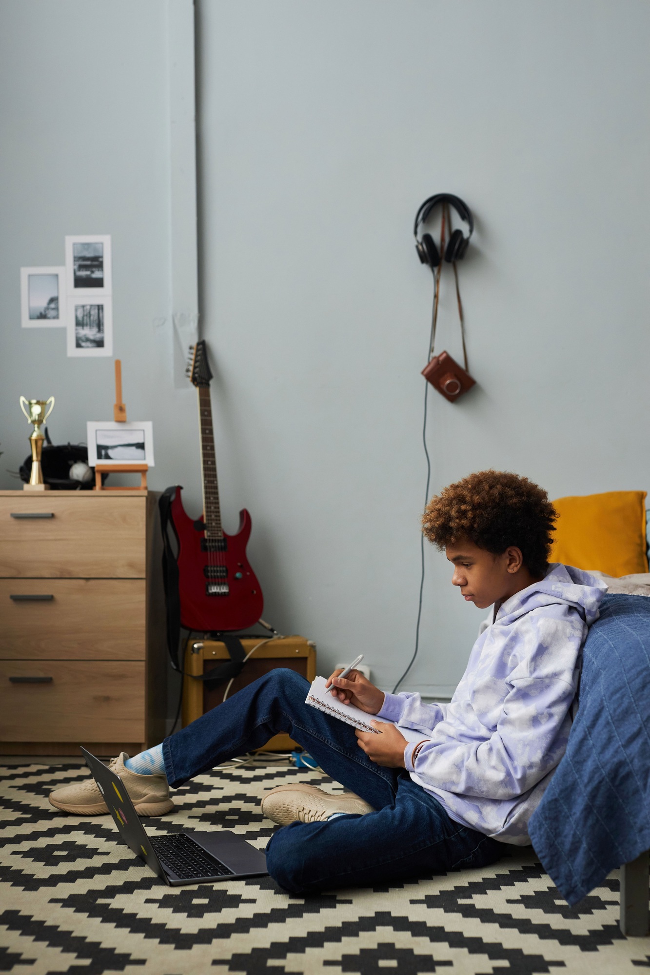 Side view of diligent schoolboy sitting in front of laptop on the floor by bed